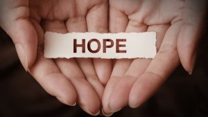 Two hands holding a piece of torn paper with the word HOPE cut out of it. 